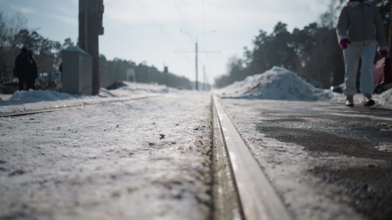 close up rail in snow along winter tram stop, low angle perspective, icy texture and sun glare, pedestrians near platform, cars parked by roadside, quiet urban mood, long shadows on pavement