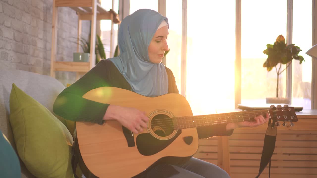 Portrait young muslim woman sitting on the couch playing guitar in a modern apartment rays of the sun