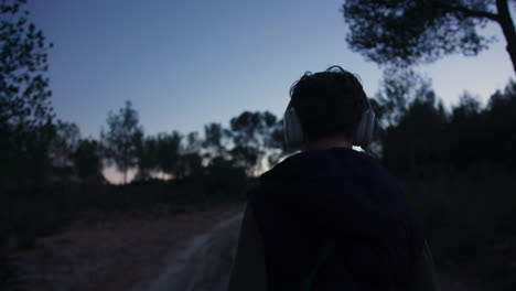 Teenage male walking rural dirt path at sunset, listening to music, headphones on, absorbing surrounding landscape serenity and natural beauty