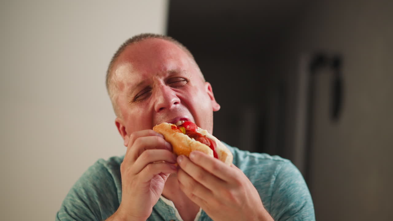 Young man seated enjoying sandwich for lunch opens mouth wide to take big bite while licking thumb, wearing casual greenish blue shirt, indoor setting with blurred background