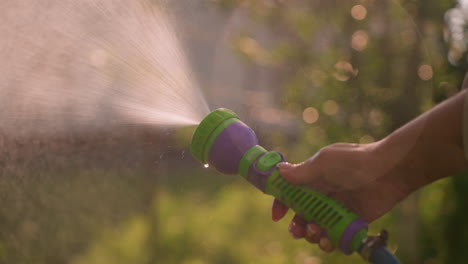 close-up of someone holding water hose spraying water outdoors, creating refreshing mist effect, with blurred background of greenery and building in soft focus