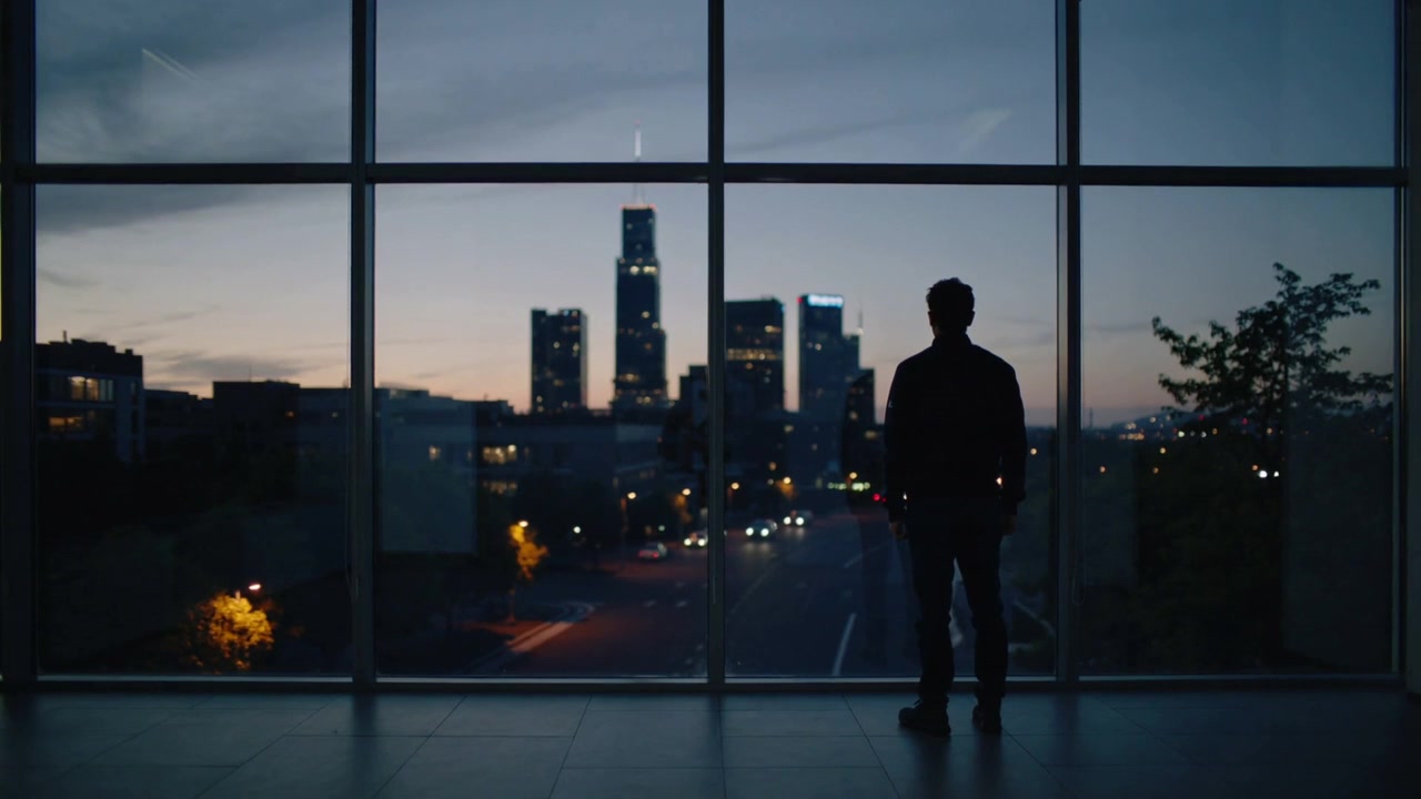 Hombre a contraluz observando el horizonte de la ciudad desde el vestíbulo, junto a una ventana de celosía, al caer la noche y notando los faros.