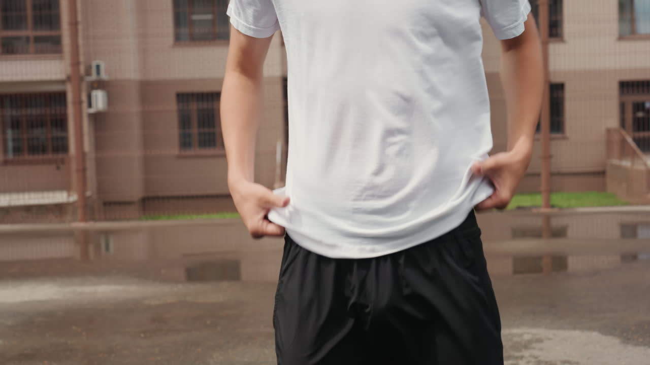 雨上がりの濡れたコートでシャツを頭上に掲げる若い男性、都会のアパートを背景に、水浸しのショートパンツとあらわになった上半身、解放感を表現したポーズ、建物が水たまりに映り込み、運動好きの雰囲気とむき出しの感情。