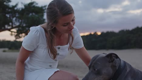 young woman petting her american staffordshire terrier dog in sand dunes and looking up to camera