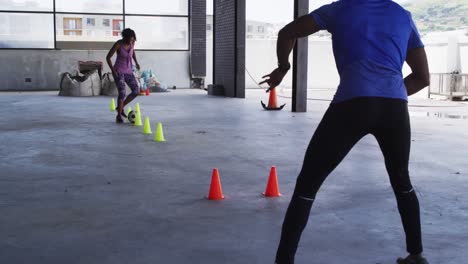 mujer afroamericana haciendo slalom con un fútbol en un edificio urbano con un hombre animándola