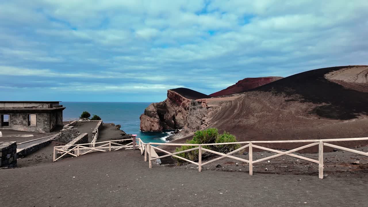 Drone footage flying toward the volcanic coastline of Capelinhos, showcasing its black sand and rugged Atlantic cliffs and the Lighthouse