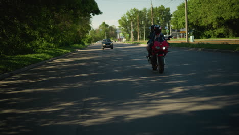dos hermanas montan una bicicleta eléctrica roja, ambas con cascos, en una carretera sombreada, se ve un coche arrancando por detrás mientras otro se mueve en el carril opuesto, los árboles alinean la carretera
