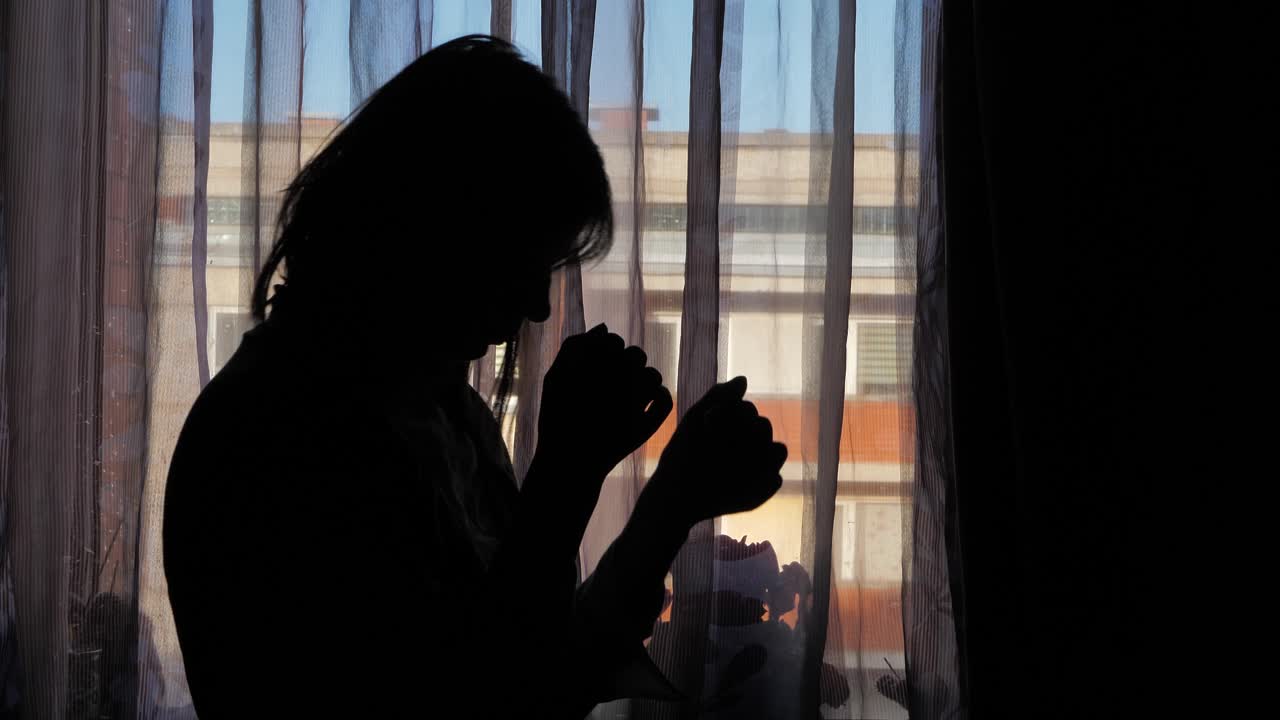A woman is seen in silhouette, head bowed near a window with sheer curtains and warm sunset light, evoking a sense of sorrow or emotional distress