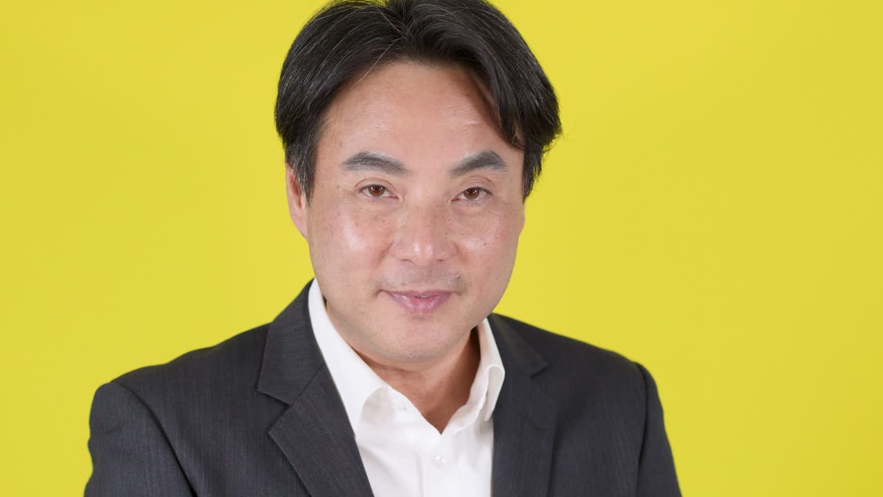 Businessman in formal suit smiles, waves hand, and gestures confidently against bright yellow backdrop