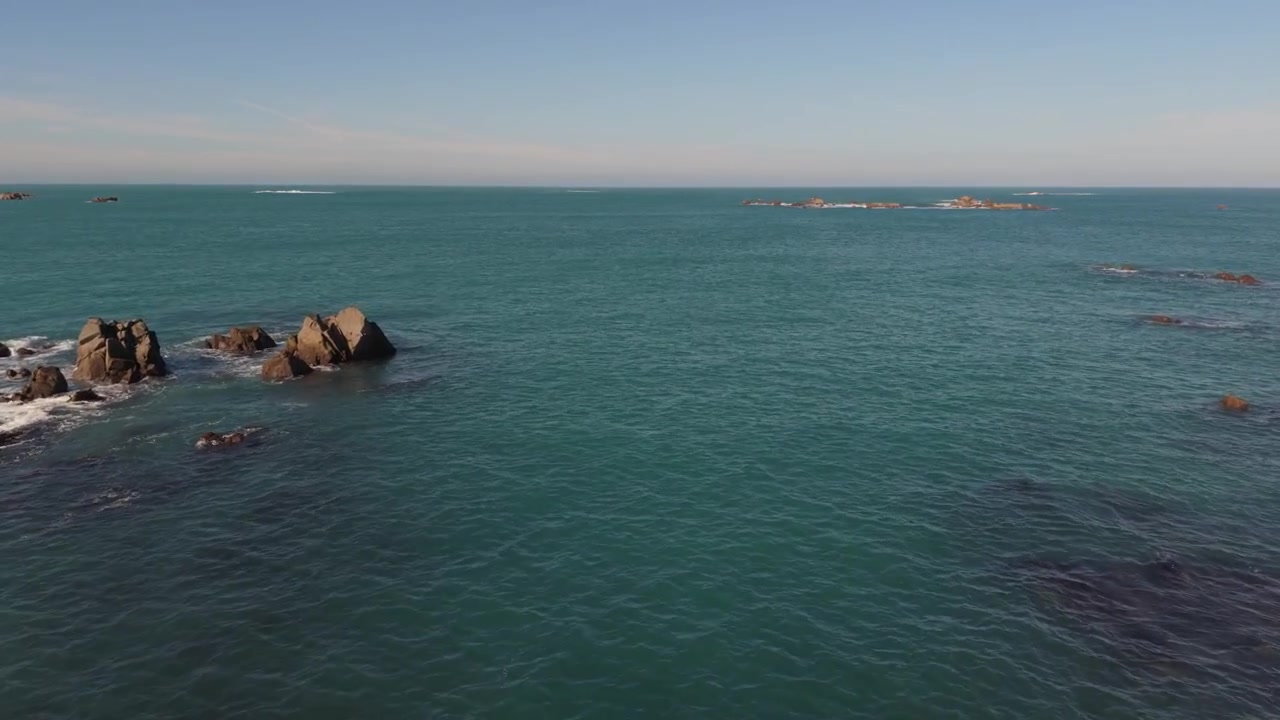 Riprese drone lente e molto basse in avanti che accelerano verso l'orizzonte su acque cristalline e calme intorno a formazioni rocciose sotto il sole del tardo pomeriggio, con onde che lambiscono dolcemente le rocce e cielo blu.