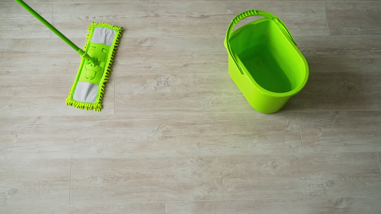 Cleaning the parquet floor with a wet mop. Green brush and a bucket on wooden floor. Housekeeper cleans the floor with a mop. Daily chores in apartment. Top view.