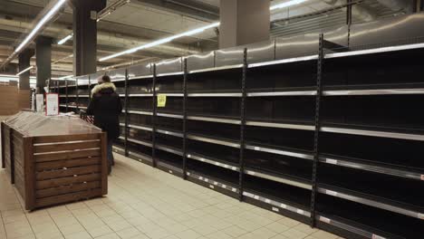 woman in warm jacket, hat walks with cart past empty shelves in russian store.