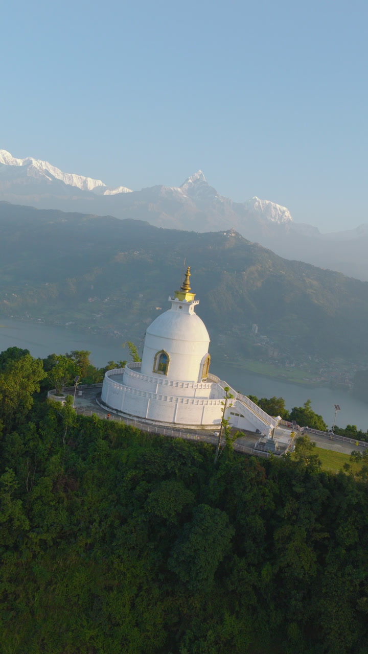 Aerial shot rotating from vertical to horizontal framing of World Peace Stupa in Pokhara Nepal surrounded by lush green forest with Annapurna peaks rising behind.