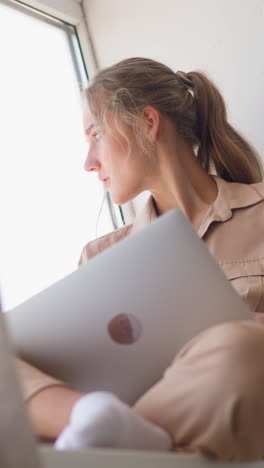 young copywriter woman looks through window to distract from working on texts on laptop behind brown curtain in comfortable room slow motion