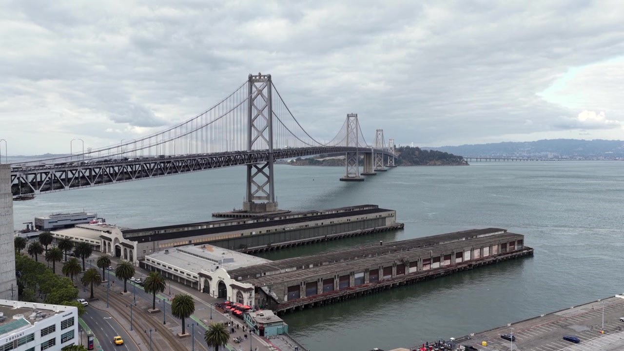 Historiske pirbygninger langs San Franciscos Embarcadero-vannfront, trafikken flyter langs palmeomkransede trikkeskinner under den ruvende Bay Bridge som strekker seg over tåkete buktvann. Luftfoto fra drone som stiger, vidvinkelbilde