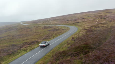 Forwards-tracking-of-vintage-silver-convertible-car-driving-on-curvy-road-surrounded-by-grasslands.-Countryside-in-remote-location.-Ireland