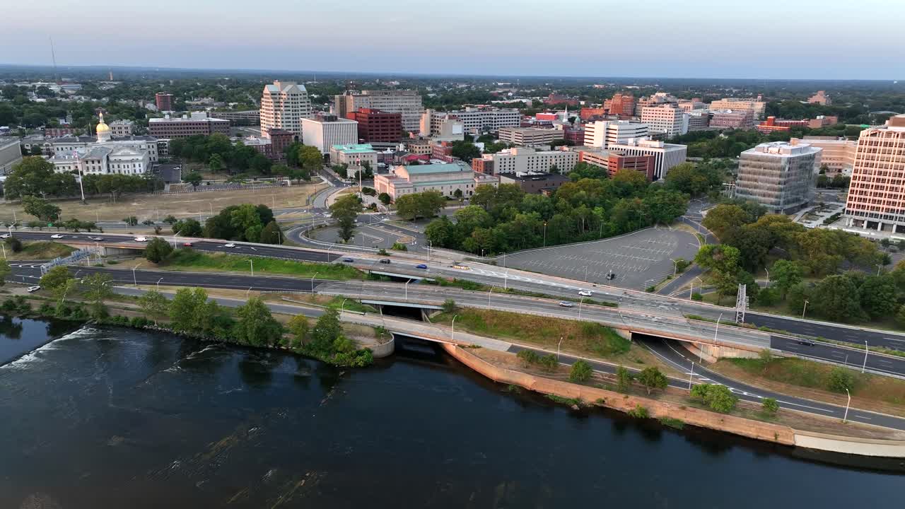 Aerial wide shot showing driving cars on interstate road in Trenton along Delaware River at sunset. Downtown with towers and blocks in background. New Jersey, USA in summer