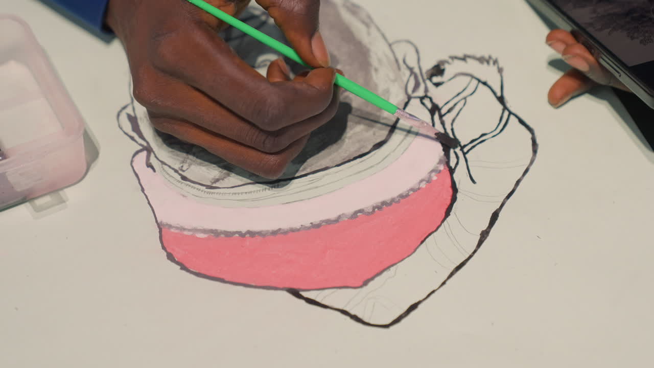Closeup portrait sketch black artist hand, careful coloring of pink hat with green brush, ink outline on white surface, small paint box nearby, focused wrist movement, soft studio light, tactile