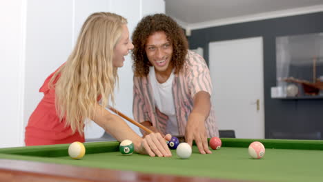 una joven caucásica y un hombre biracial disfrutan de un juego de billar en casa.