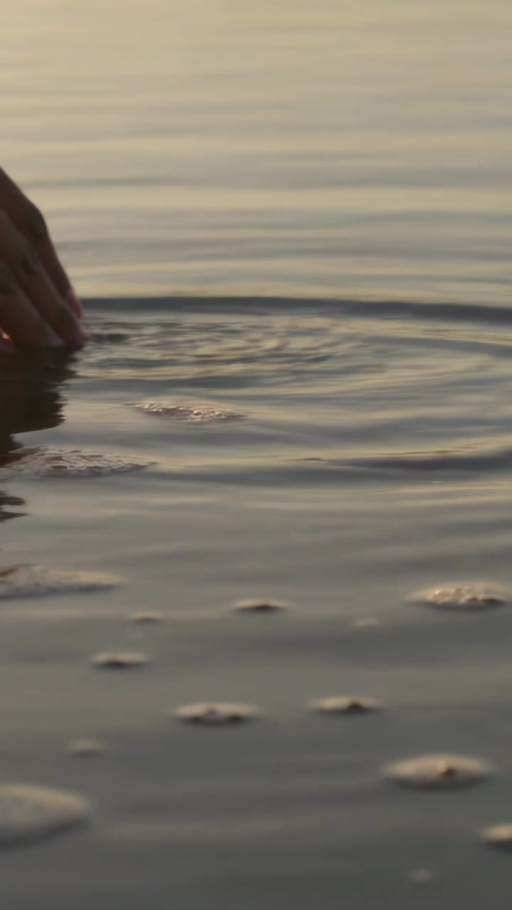 Video verticale: Mano che si avvicina e tocca l'acqua bassa della riva, formando increspature e schiuma, spazio per il testo