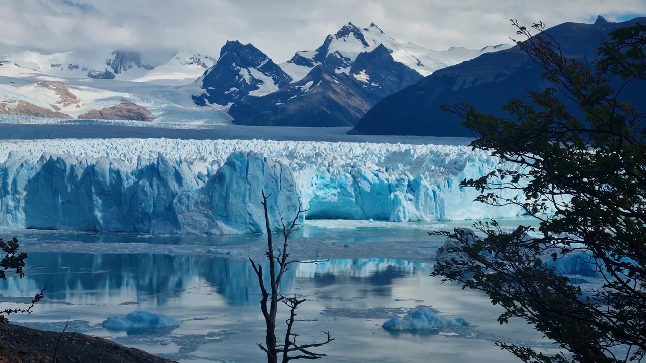 Il Ghiacciaio Perito Moreno in Patagonia, Argentina, si erge come un'immensa parete di ghiaccio blu sopra il Lago Argentino, riflettendosi sotto le cime innevate delle Ande in una giornata nuvolosa e suggestiva.