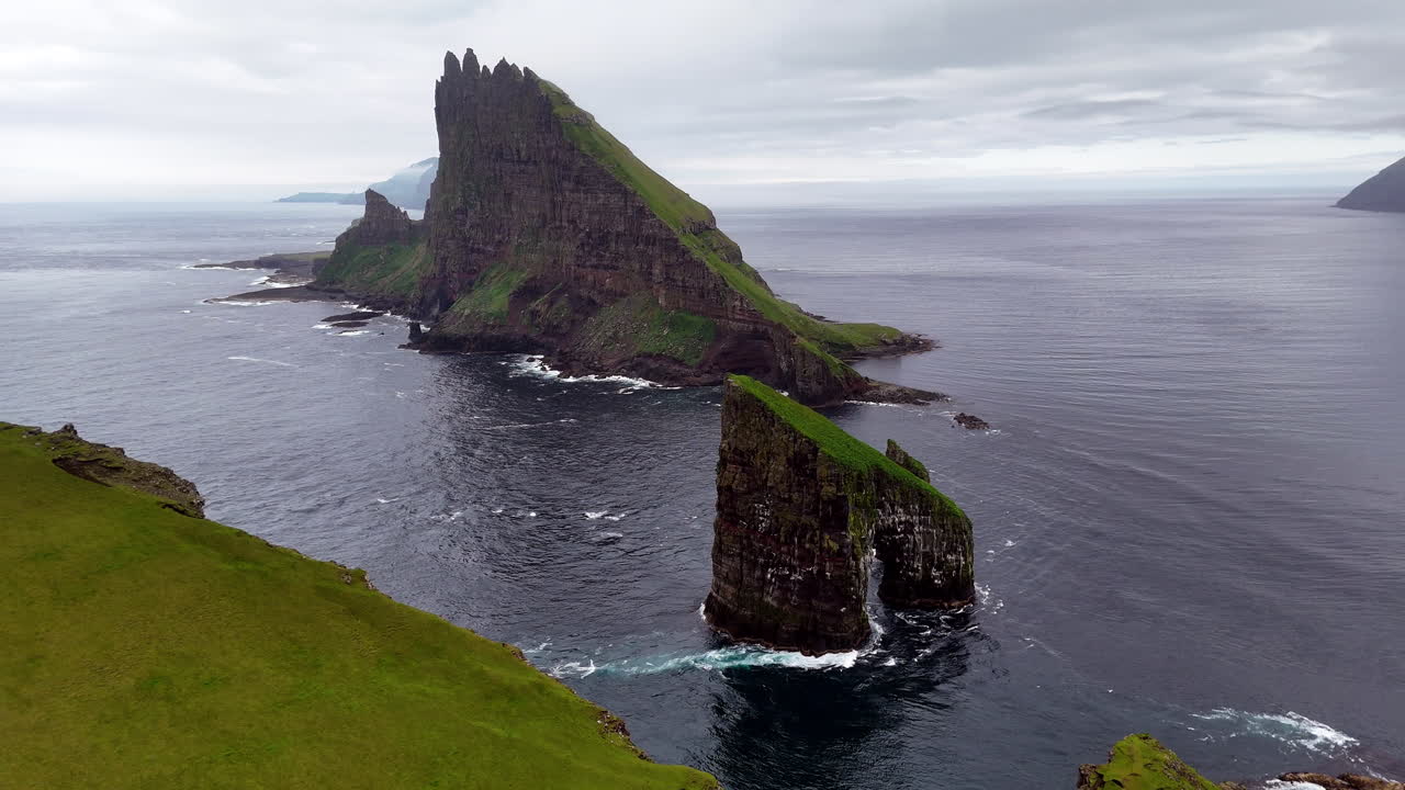 Cinematic aerial view of Drangarnir sea stacks rising dramatically from the Atlantic Ocean near Vágar, Faroe Islands, showcasing rugged cliffs, lush green slopes, and misty Nordic seascape