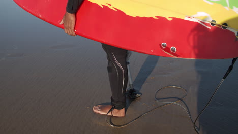 surfista maschio serio in muta con gamba artificiale in piedi sulla riva dell'oceano e tenendo la tavola da surf sotto il braccio