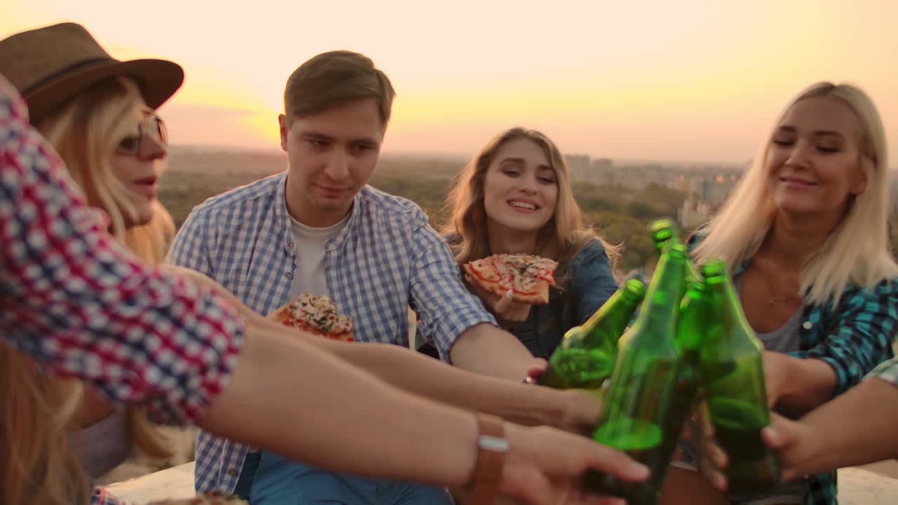 Young men and women clinks glasses and drinks beer from green bottels on the party with friends on the roof at the sunset.They eat hot pizza after in summer everning.