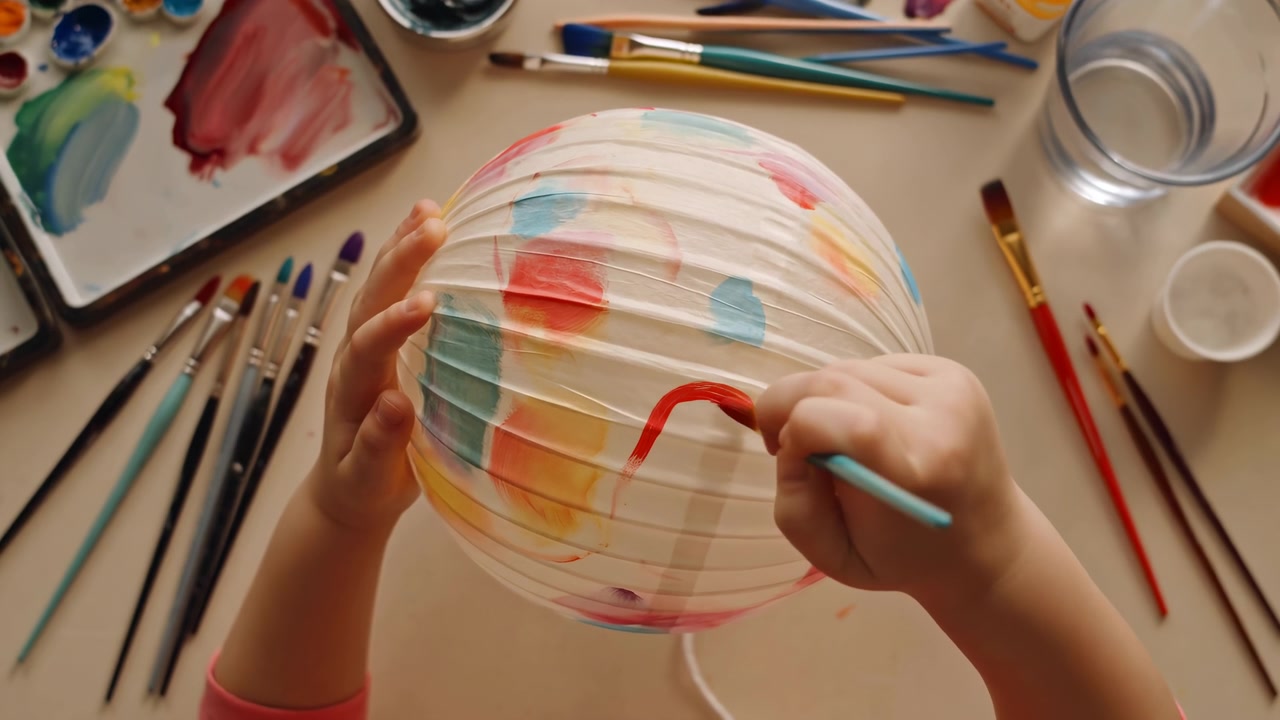 Close overhead oblique view of child's hands painting round pleated paper lantern with small round brush, adding short curved red stroke over blue, yellow, and green swatches on tabletop during