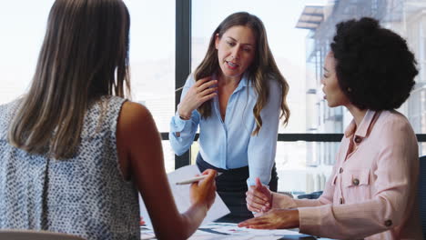 female multi-cultural business team meet around boardroom table with laptops discussing documents
