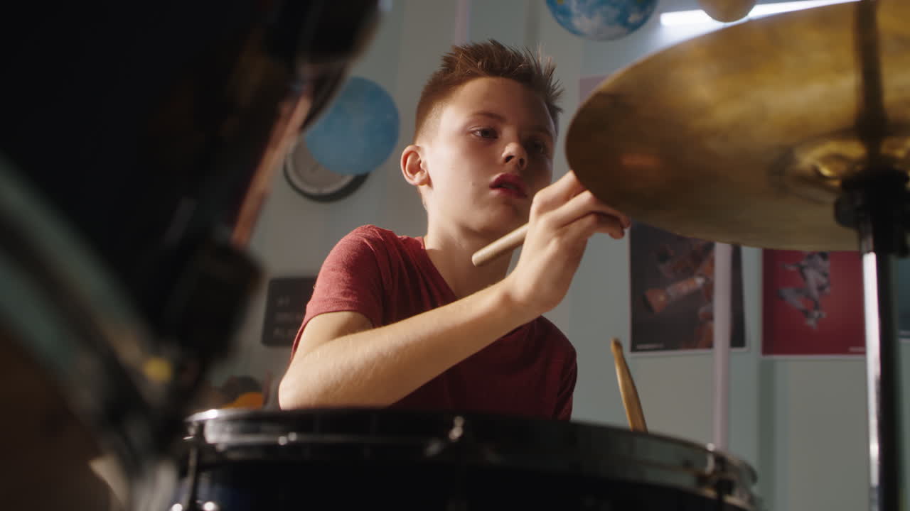 Teenager Playing Drums at Home from below serious Teen Musician in Red T Shirt Practicing to Play Drums during Music Session at Home