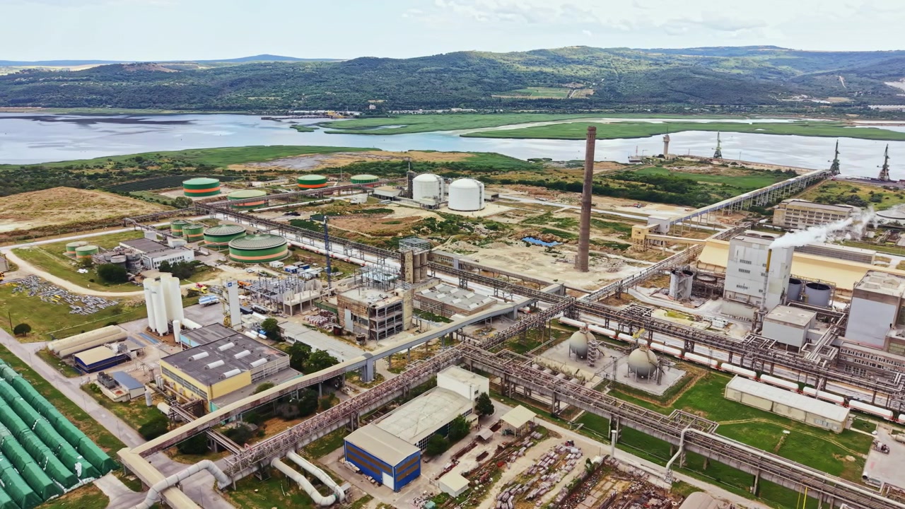 Complejo industrial situado cerca del agua con grandes tanques de almacenamiento y tuberías en un paisaje verde bajo un cielo nublado.