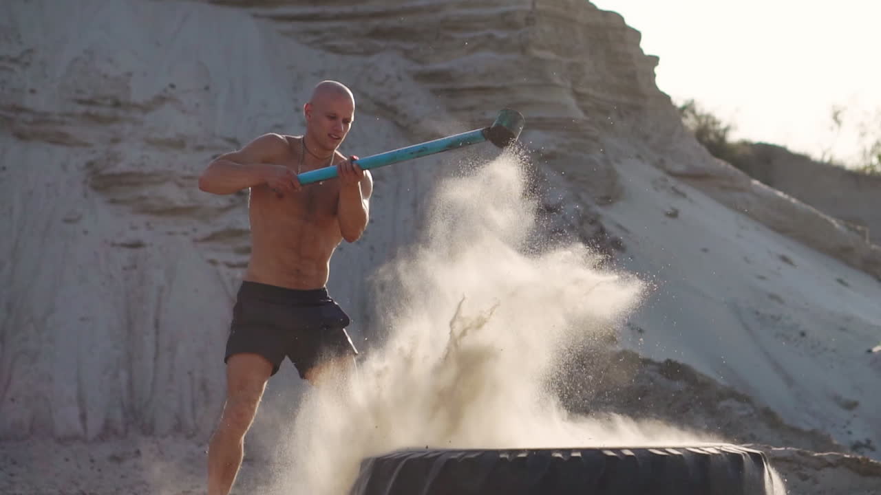 ⁇ 머리 강인한 남자가 모래 산에서 거대한 바 ⁇ 에 망치를 쳐서 느린 영상으로 힘과 지구력 훈련을 합니다.