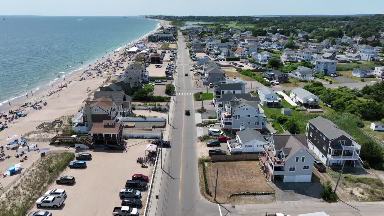 Inseguimento aereo di un'auto sulla strada costiera principale di Misquamicut, Rhode Island. Giornata estiva soleggiata con spiaggia sabbiosa affollata e acqua turchese dell'Oceano Atlantico. Campo lungo. Case sulla spiaggia e abitazioni negli USA