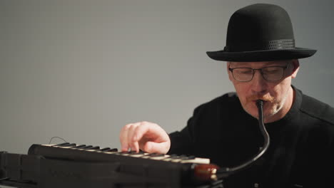 A keyboardist wearing a black hat,glasses, and a black outfit sits and plays a red sampler keyboard against a grey background. The musician is focused intently on the performance
