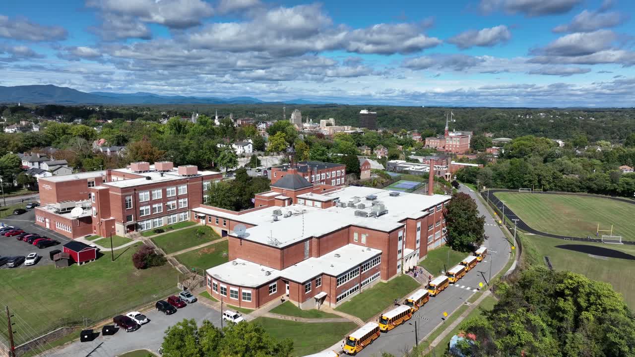 American school scene with row of waiting yellow school buses in front of historic red brick buildings. Aerial descend wide shot. Sunny day in autumn season. Pennsylvania, USA