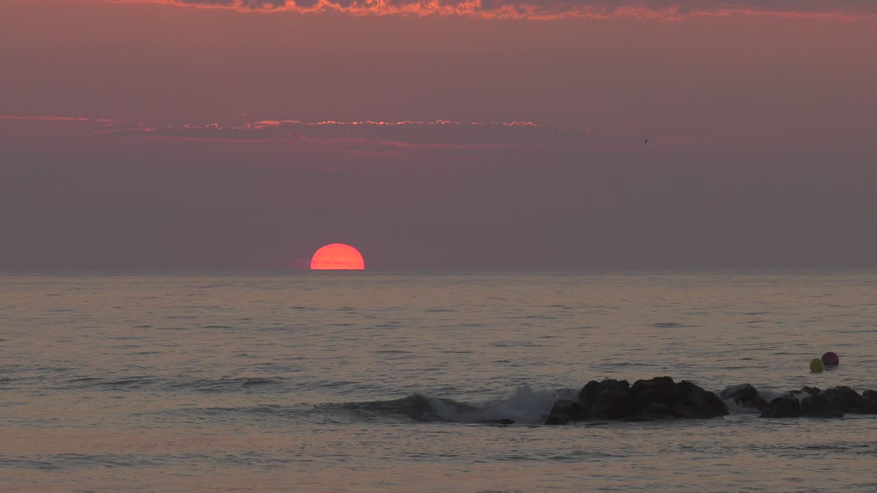 Orange sunrise over ocean horizon with calm sea, waves breaking on rocks