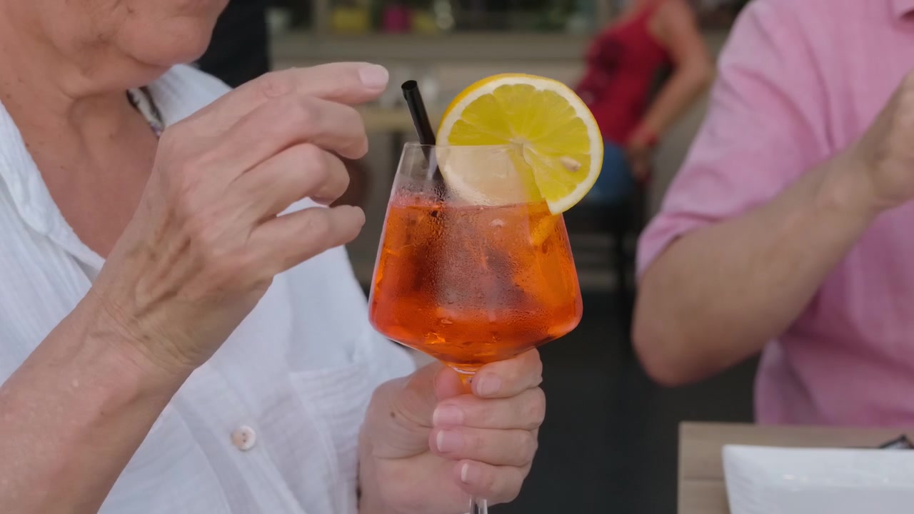 Primo piano di una donna anziana che beve un cocktail spritz all'arancia con una cannuccia.