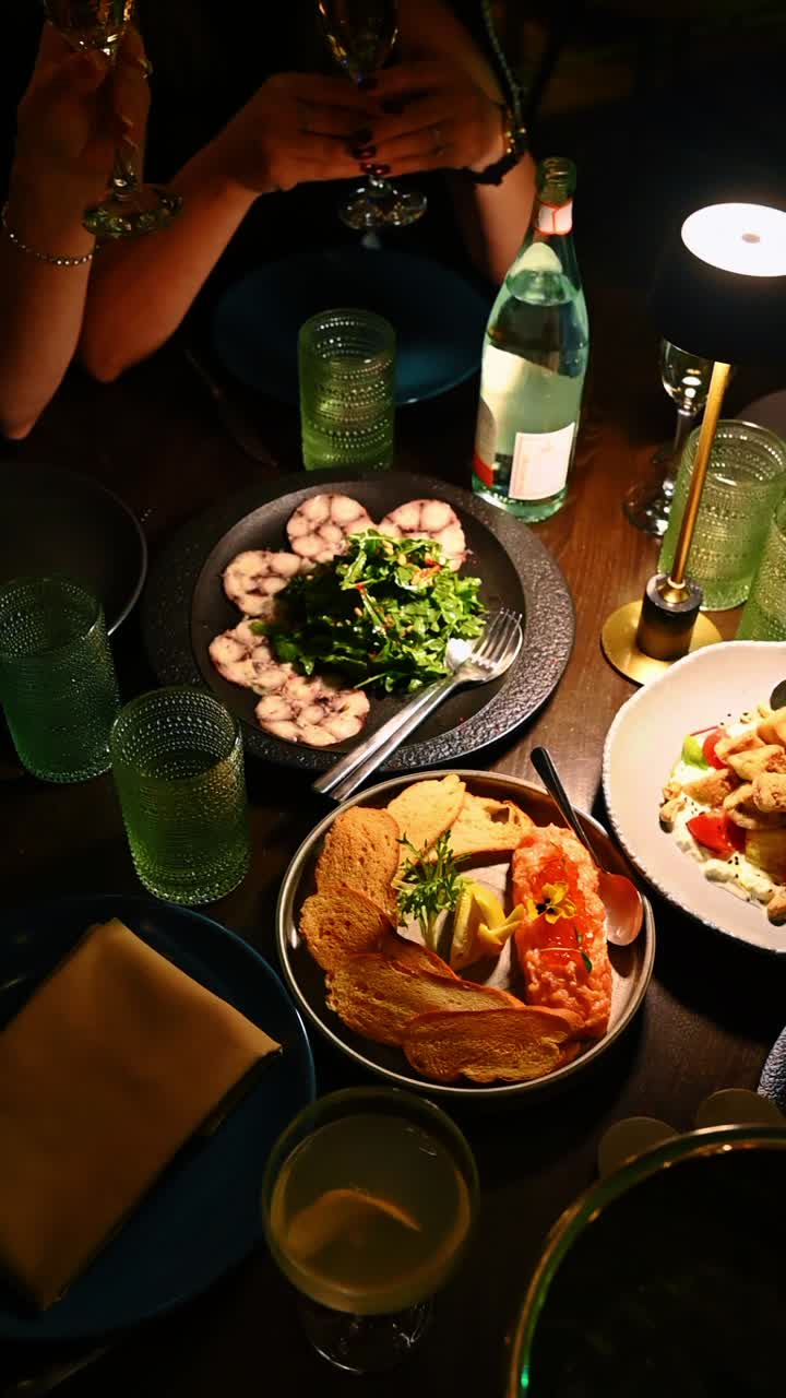 Gourmet dinner table with octopus carpaccio and salmon tartare. Top view of restaurant table featuring seafood dishes, green glassware and people toasting with wine glasses