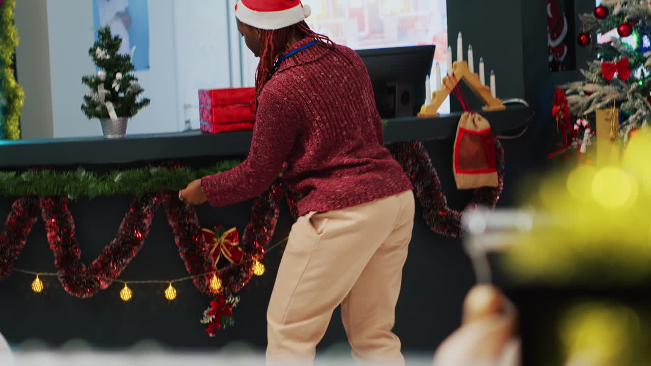 empleado afroamericano con sombrero de santa decorando una tienda de ropa con adornos de navidad antes de un evento promocional festivo. trabajador instalando un árbol de navidad en una tienda de moda, ayudado por un colega
