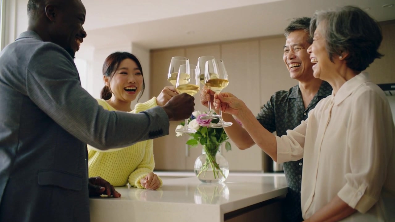 Four people raising wine glasses around kitchen island after toast prompt, with vase, in jacket