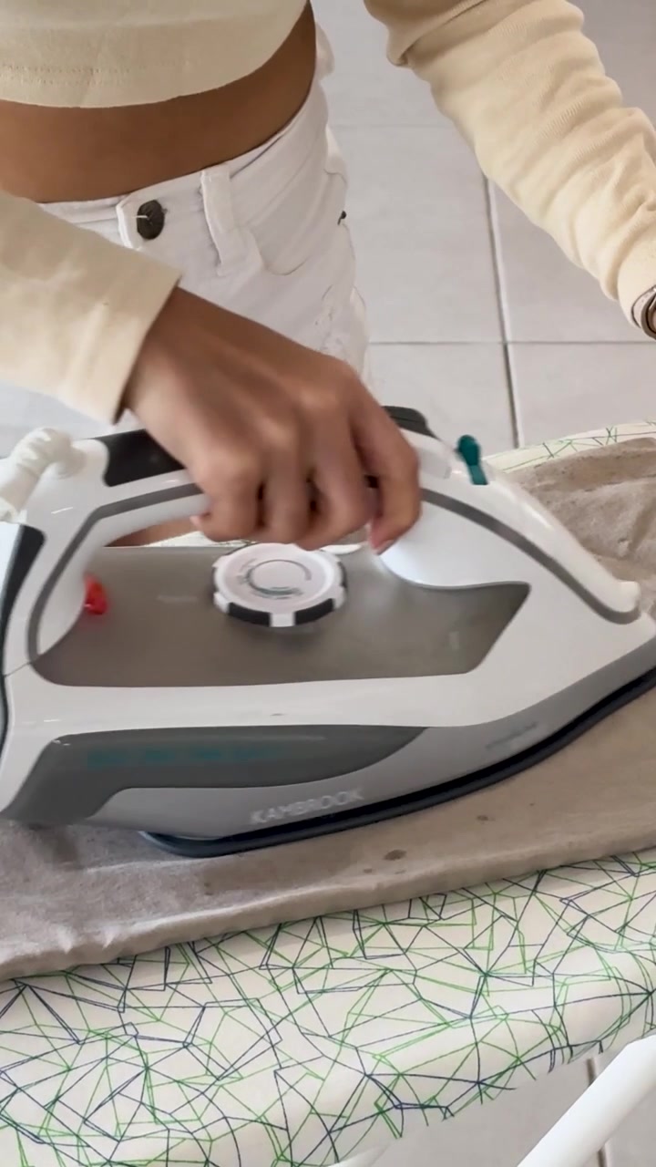A woman uses a steam iron on khaki trousers over a patterned ironing board.