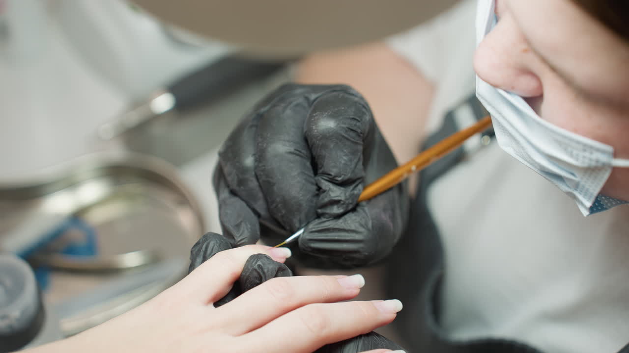 Close-up of nail technician wearing black glove and face mask carefully using fine brush to fill nail polish evenly along nail surface during detailed manicure session in professional salon setting