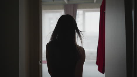 back view female trainer walking into room through open door with red garments hanging by window and purple curtains under soft natural light capturing serene fitness environment transition