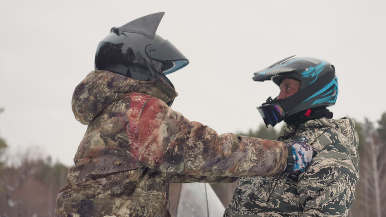Snowmobile riders check helmets snowy clearing, two friends in camo jackets adjust chin straps, tense supportive exchange, forest background, overcast sky, hands on shoulders, preride safety moment