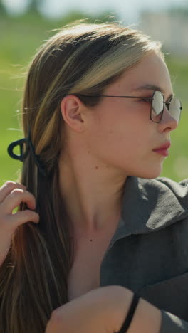 dama con una camiseta negra debajo de una chaqueta gris y gafas de sol, ajustando su cabello con una expresión enfocada, el escenario es al aire libre, con vegetación borrosa en el fondo en un día brillante