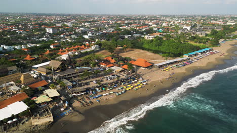 ascending top down shot of crowded surfer beach with crashing waves on bali island -rising drone shot with cityscape view