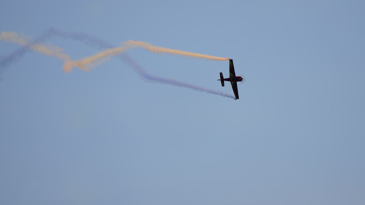 Acrobatic airplane leaves blue and yellow trail of smoke while flying upward against clear blue sky