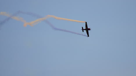 acrobatic airplane leaves blue and yellow trail of smoke while flying upward against clear blue sky