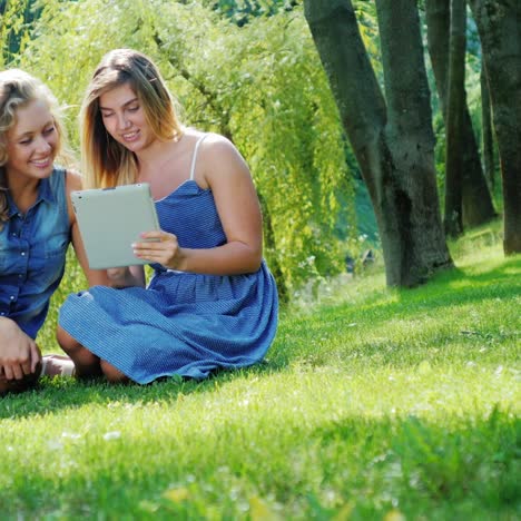 Two-young-women-relax-in-the-park-and-use-a-tablet